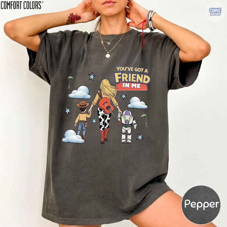 May include: A charcoal gray short-sleeve t-shirt featuring a graphic of a person with blonde hair holding hands with a cowboy and a space ranger. The text reads YOU'VE GOT A FRIEND IN ME. The design includes clouds, stars, and a guitar.