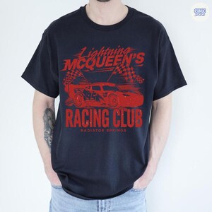 May include: Navy blue t-shirt featuring a red graphic of Lightning McQueen's Racing Club. The design includes a race car, checkered flags, and the text "Lightning McQueen's Racing Club, Radiator Springs."