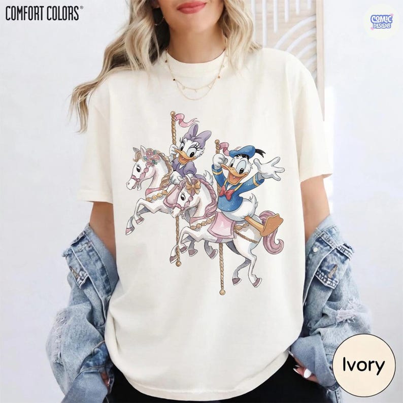 May include: Ivory-colored t-shirt featuring a carousel scene with Donald Duck and Daisy Duck riding white horses. The design includes pink and gold accents, with the "COMFORT COLORS" and "COMIC DESIGNS" logos visible. The shirt is a crew neck.