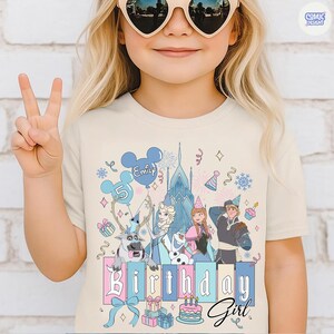 May include: A beige t-shirt featuring a colorful graphic of characters from a popular animated movie, including a castle, reindeer, and birthday elements. The text "Birthday Girl" is prominently displayed.