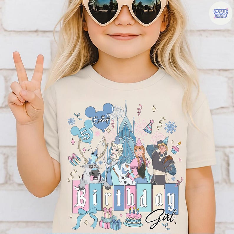 May include: A beige t-shirt featuring a colorful graphic of characters from a popular animated movie, including a castle, reindeer, and birthday elements. The text "Birthday Girl" is prominently displayed.