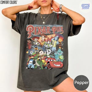 Pu&ograve; includere: T-shirt color pepe con grafica dei personaggi di Pixar Pier. Il design include personaggi di Toy Story, Monsters & Co. e Cars. Il testo "PIXAR PIER" &egrave; visualizzato in un font in stile vintage.