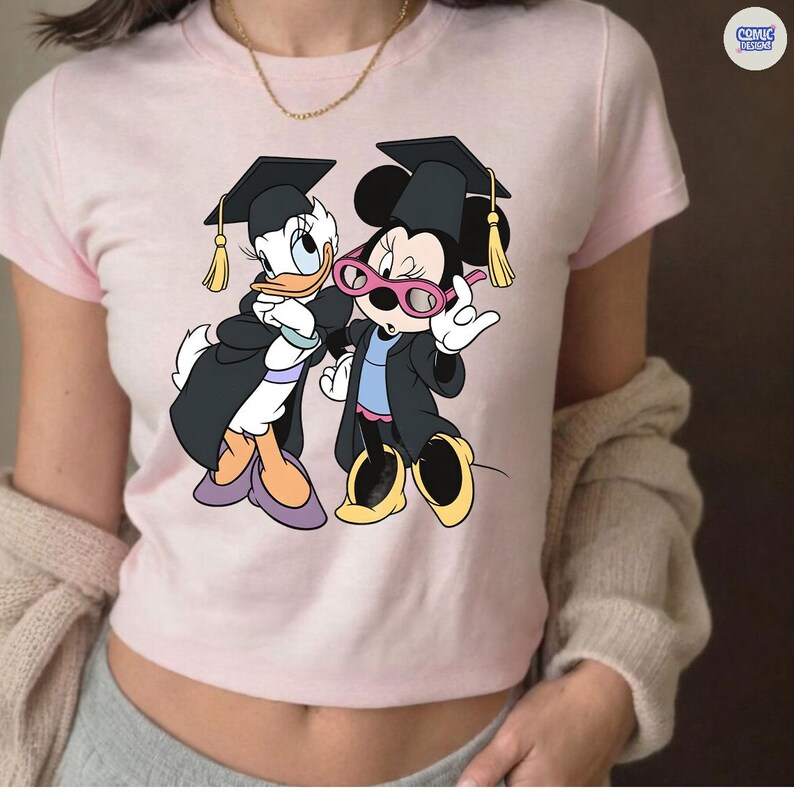 Puede incluir: Camiseta rosa claro con un dise&ntilde;o de dibujos animados de Daisy y Minnie Mouse con atuendos de graduaci&oacute;n. Daisy y Minnie Mouse llevan vestidos y birretes negros con borlas doradas. El logotipo de Comic Designs es visible en la esquina superior derecha.