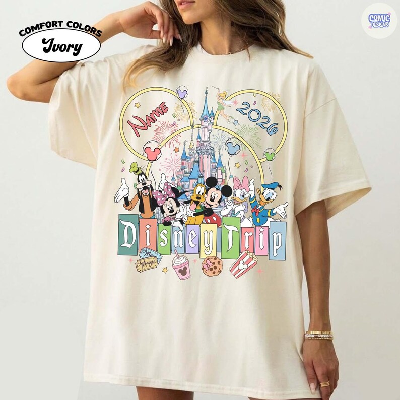 May include: Ivory Comfort Colors t-shirt featuring a colorful Disney Trip design. The design includes Mickey Mouse, Minnie Mouse, Goofy, Donald Duck, and Daisy Duck, with a castle and fireworks. The text "Name" and "2026" are also included.