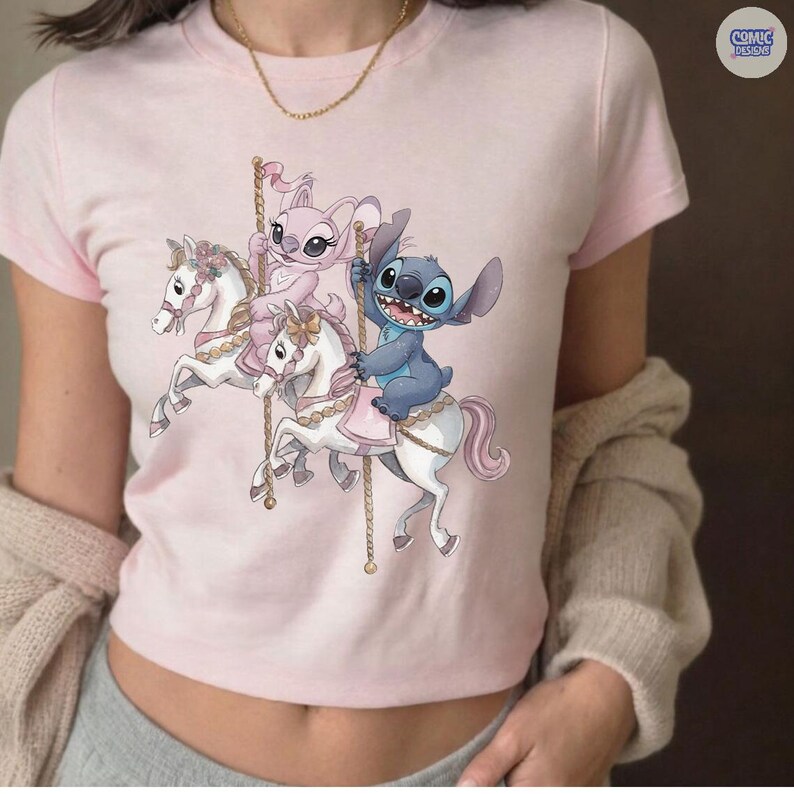 May include: A light pink t-shirt featuring a carousel design with the characters Stitch and Angel. Stitch is riding a white horse, and Angel is riding another. The shirt has a crew neck and short sleeves. The text "COMIC DESIGNS" is in the upper right corner.