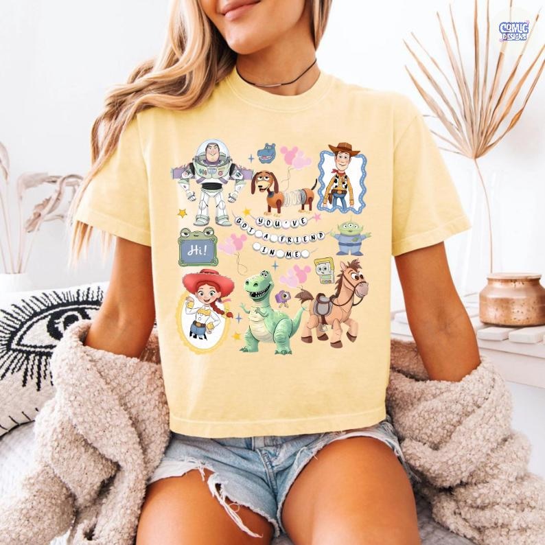 May include: A pale yellow t-shirt featuring a colorful graphic print of various Toy Story characters including Buzz Lightyear, Woody, Jessie, Rex, Slinky Dog, and Bullseye, with the text "YOU'VE GOT A FRIEND IN ME" arranged in a circular banner.
