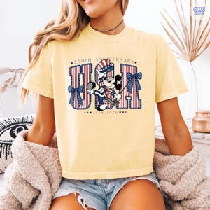 Op de afbeelding: Een lichtgeel t-shirt met een afbeelding van Mickey Mouse die een patriottische hoge hoed draagt. De tekst luidt 250TH ANNIVERSARY USA 1776 2026. De letters U, S en A zijn gevuld met rode en witte verticale strepen en versierd met blauwe strikken.