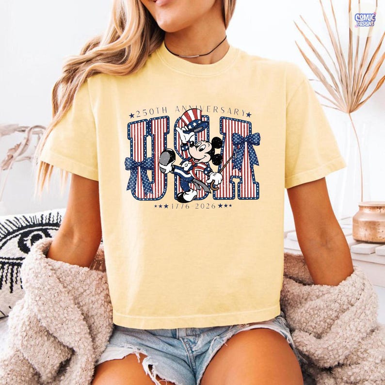 Op de afbeelding: Een lichtgeel t-shirt met een afbeelding van Mickey Mouse die een patriottische hoge hoed draagt. De tekst luidt 250TH ANNIVERSARY USA 1776 2026. De letters U, S en A zijn gevuld met rode en witte verticale strepen en versierd met blauwe strikken.