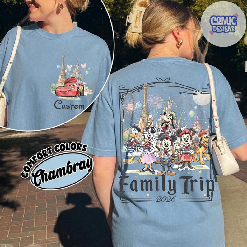 May include: A light blue t-shirt featuring a small graphic of a red race car and a castle on the front left chest. The back displays a large illustration of cartoon characters in front of a castle and Eiffel Tower with the text Family Trip 2026.