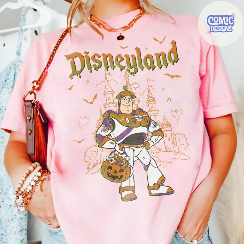 Może przedstawiać: Jasnor&oacute;żowy t-shirt z grafiką Buzza Astrala trzymającego wiaderko w kształcie dyni na tle zarysu zamku. Napis "Disneyland" jest wydrukowany zieloną, ściekającą czcionką nad postacią, w towarzystwie małych nietoperzy i duch&oacute;w.