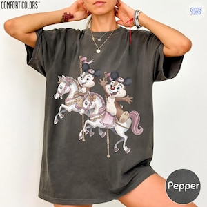 May include: Dark gray Comfort Colors t-shirt featuring a carousel design with cartoon characters. The carousel horses are white with pink accents, and the characters are in the center. The shirt has the color name "Pepper" on the bottom right.