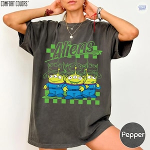 May include: A charcoal gray short-sleeve t-shirt featuring a graphic print of three green three-eyed aliens in blue space suits. The design includes the word Aliens in green cursive text above the characters, set against a green checkered background.