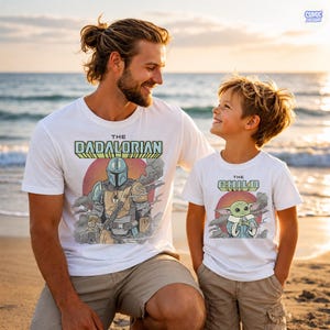Puede incluir: Camisetas blancas con gr&aacute;ficos a juego. Una camiseta dice "The Dadalorian" con una ilustraci&oacute;n de Mandaloriano. La otra dice "The Child" con una ilustraci&oacute;n de Grogu. Ambas camisetas tienen un dise&ntilde;o retro.