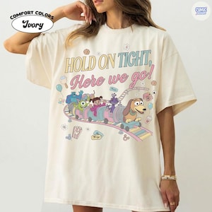 May include: Ivory Comfort Colors t-shirt featuring a colorful graphic of a cartoon train with characters and the text "HOLD ON TIGHT, Here we go!". The design includes a dog, monsters, and other animated figures on a roller coaster.
