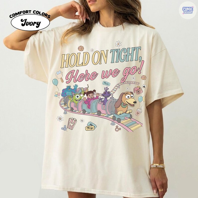 May include: Ivory Comfort Colors t-shirt featuring a colorful graphic of a cartoon train with characters and the text "HOLD ON TIGHT, Here we go!". The design includes a dog, monsters, and other animated figures on a roller coaster.