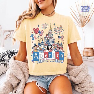 May include: A pale yellow t-shirt featuring a graphic of a castle, fireworks, and cartoon characters wearing patriotic red, white, and blue accessories. The text reads America, 250 Years of Freedom, and 1776-2026, with balloons and stars.