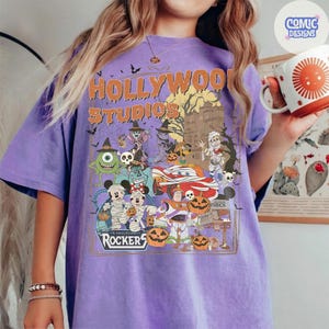 K&ouml;nnte beinhalten: Ein violettes T-Shirt mit einem Grafikdruck, der den Text Hollywood Studios und Rockers enth&auml;lt. Das Design zeigt verschiedene Zeichentrickfiguren in Halloween-Kost&uuml;men, K&uuml;rbisse, Flederm&auml;use und die Silhouette eines Spukhauses auf violettem Stoff.