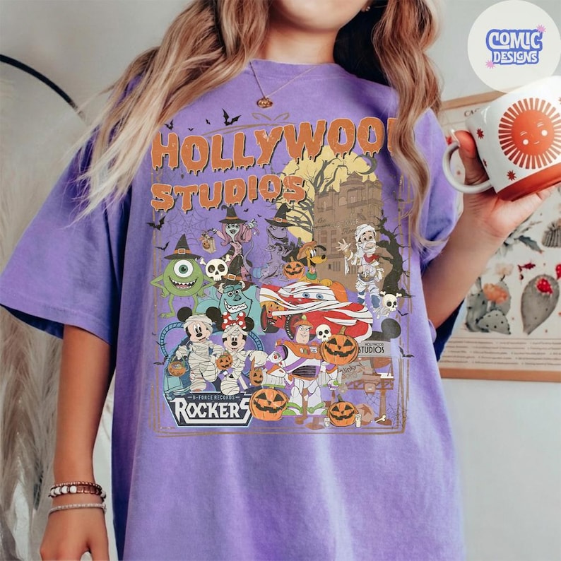 K&ouml;nnte beinhalten: Ein violettes T-Shirt mit einem Grafikdruck, der den Text Hollywood Studios und Rockers enth&auml;lt. Das Design zeigt verschiedene Zeichentrickfiguren in Halloween-Kost&uuml;men, K&uuml;rbisse, Flederm&auml;use und die Silhouette eines Spukhauses auf violettem Stoff.