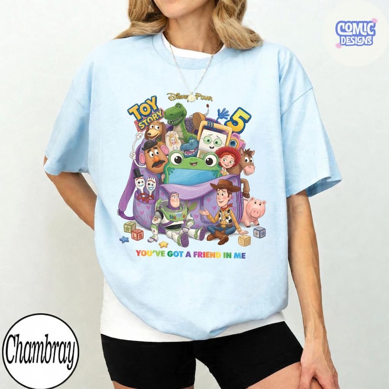 May include: A light blue t-shirt featuring a colourful graphic of Toy Story characters including Woody, Buzz Lightyear, and Rex emerging from a purple backpack. The text reads Toy Story 5 and You've Got A Friend In Me. A small logo in the corner says Comic Designs.