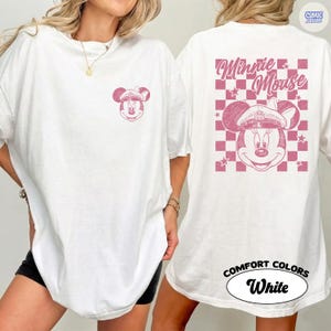 May include: White t-shirt with a pink Minnie Mouse graphic. The front has a small Minnie Mouse design, and the back features a larger design with the words "Minnie Mouse" and a pink and white checkered pattern. The shirt is labelled "Comfort Colors White."
