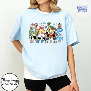 May include: A light blue short-sleeve t-shirt featuring a graphic print of the word NAME filled with various animated characters from the Toy Story franchise, including Woody, Buzz Lightyear, Jessie, Rex, and Slinky Dog, with a small logo in the corner.