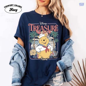 Op de afbeelding: Marineblauw Comfort Colors T-shirt met een Disney Winnie the Pooh ontwerp. De afbeelding bevat het woord "TREASURE" en een cartoon Winnie de Poeh in een matrozenoutfit, met palmbomen en een boot op de achtergrond.