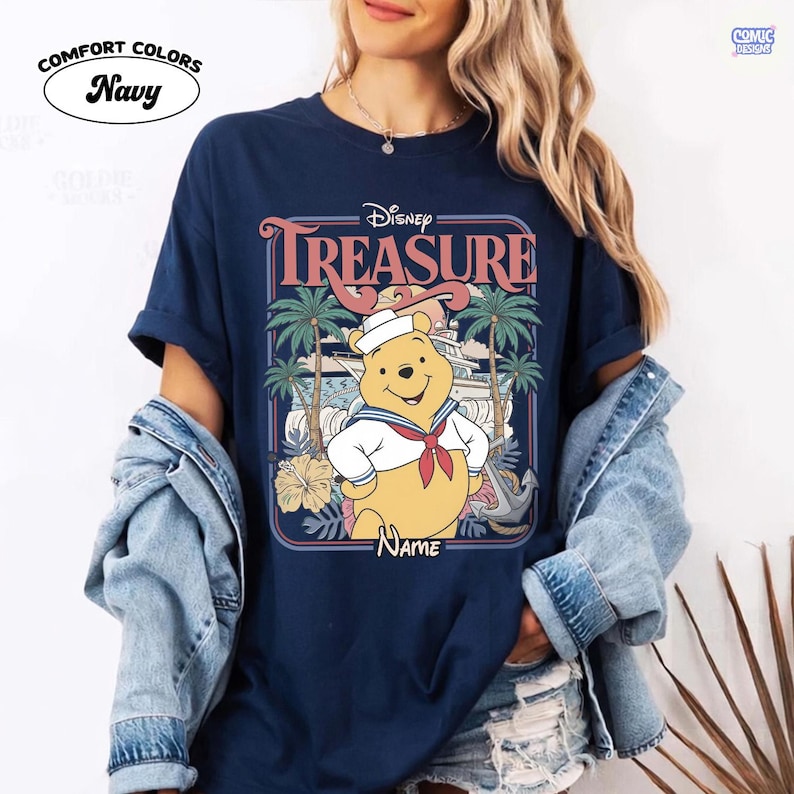 Op de afbeelding: Marineblauw Comfort Colors T-shirt met een Disney Winnie the Pooh ontwerp. De afbeelding bevat het woord "TREASURE" en een cartoon Winnie de Poeh in een matrozenoutfit, met palmbomen en een boot op de achtergrond.