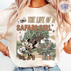 May include: Cream-colored t-shirt featuring a safari-themed graphic. The design includes the text "THE LIFE OF A SAFARI GIRL" with illustrations of Minnie Mouse and Daisy Duck in safari attire, a jeep, and other safari elements.