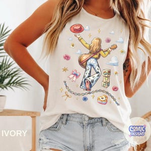 May include: An ivory sleeveless tank top featuring a graphic illustration of a person with long blonde hair playing a guitar, wearing a red hat and cow-print bell-bottom pants, surrounded by flowers, clouds, stars, and a beaded string with text.