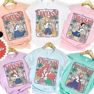 Op de afbeelding: Een verzameling pastelkleurige t-shirts met Disney-prinsessenontwerpen. Elk shirt toont een prinsessenillustratie in een rechthoekige lijst met de woorden "Fantasy", "Treasure", "Wish", "Dream" en "Adventure".