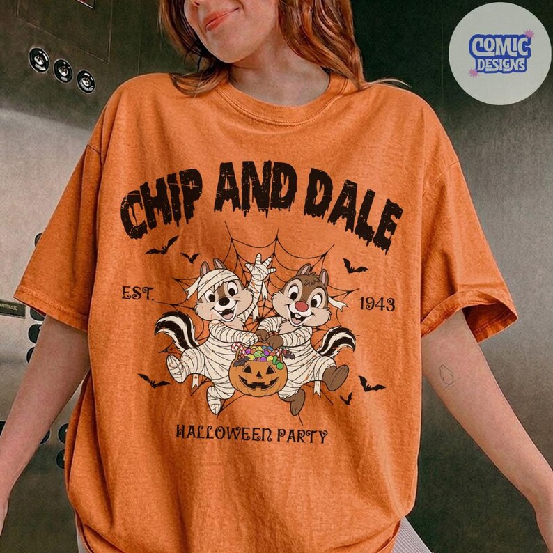 May include: An orange t-shirt featuring a graphic of Chip and Dale dressed as mummies holding a jack-o'-lantern. The text reads CHIP AND DALE, EST. 1943, and HALLOWEEN PARTY, surrounded by spiderwebs and bats. The design has a distressed, vintage look.