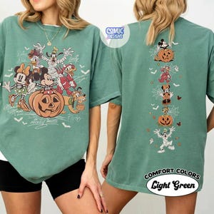 Op de afbeelding: Een saliegroen t-shirt met een grote afbeelding op de voorkant van stripfiguren in Halloween-kostuums met een pompoen en witte tekst. De achterkant toont een verticale kolom van dezelfde figuren met pompoenen, spinnenwebben en kleine geesten.