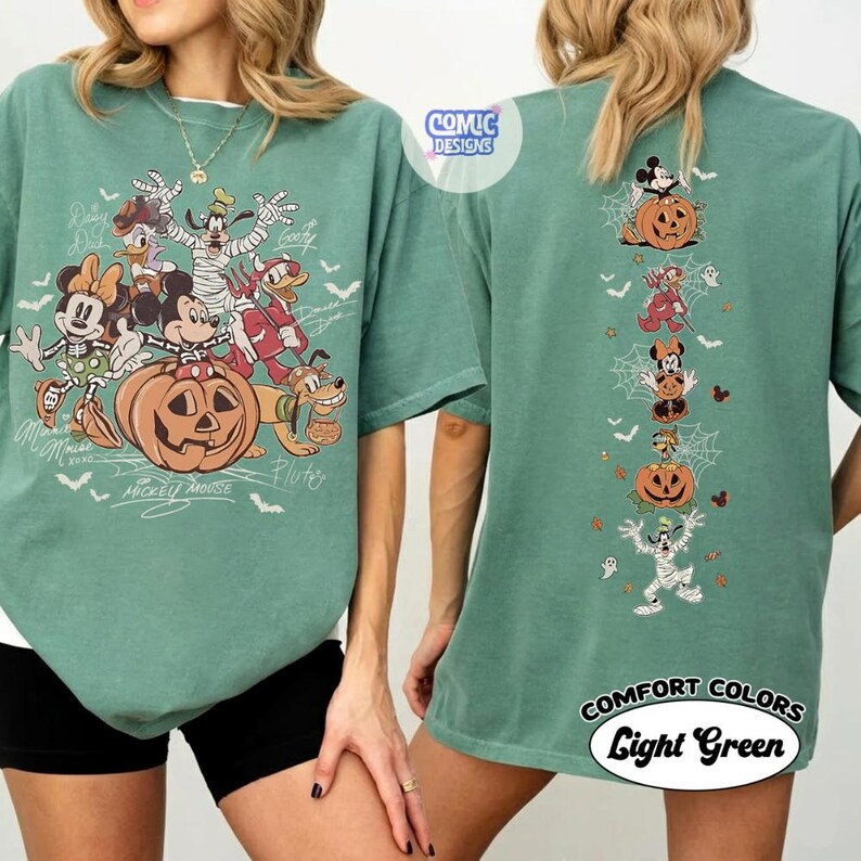 Op de afbeelding: Een saliegroen t-shirt met een grote afbeelding op de voorkant van stripfiguren in Halloween-kostuums met een pompoen en witte tekst. De achterkant toont een verticale kolom van dezelfde figuren met pompoenen, spinnenwebben en kleine geesten.