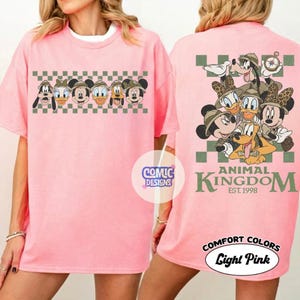 Pode incluir: T-shirt rosa claro com um design de desenho animado de personagens da Disney. A frente exibe rostos de personagens com chap&eacute;us de safari acima de um padr&atilde;o xadrez. A parte de tr&aacute;s apresenta um grupo de personagens com o texto "ANIMAL KINGDOM EST. 1998".