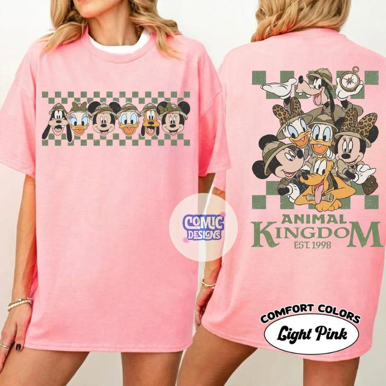 Pode incluir: T-shirt rosa claro com um design de desenho animado de personagens da Disney. A frente exibe rostos de personagens com chap&eacute;us de safari acima de um padr&atilde;o xadrez. A parte de tr&aacute;s apresenta um grupo de personagens com o texto "ANIMAL KINGDOM EST. 1998".