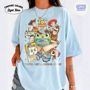 May include: A light blue t-shirt featuring a Toy Story 5 graphic with characters including Woody, Buzz Lightyear, Jessie, Bullseye, and others surrounding a cardboard box with the name Jason printed on it and the text You've Got A Friend In Me.