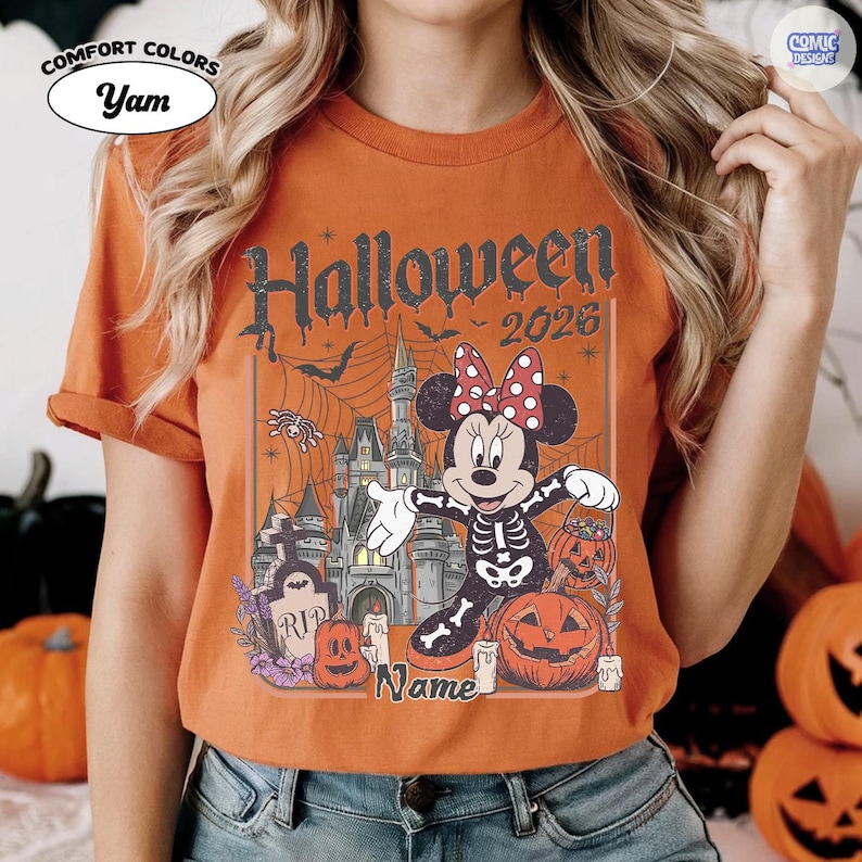 May include: Orange t-shirt with a Halloween graphic. The design includes Minnie Mouse in a skeleton costume, a haunted castle, pumpkins, and the text "Halloween 2026". The shirt has the Comfort Colors and Comic Designs logos.