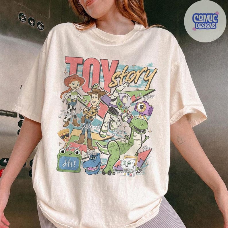 May include: A cream-colored short-sleeve t-shirt featuring a colorful graphic print of Toy Story characters including Woody, Jessie, Buzz Lightyear, Rex, and others. The design includes the text Toy Story and various small icons like a pizza slice.