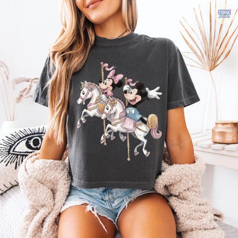 May include: Dark gray t-shirt featuring a carousel scene with Mickey and Minnie Mouse. The carousel horses are white with pink and gold accents. Mickey Mouse is wearing blue shorts and a yellow hat. The text "Comic Designs" is in the upper right corner.