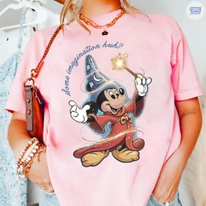 May include: Pale pink t-shirt with a graphic of Mickey Mouse as a wizard, holding a wand with a star. The text "Some imagination huh?" is above the character. The shirt has short sleeves and a round neck.