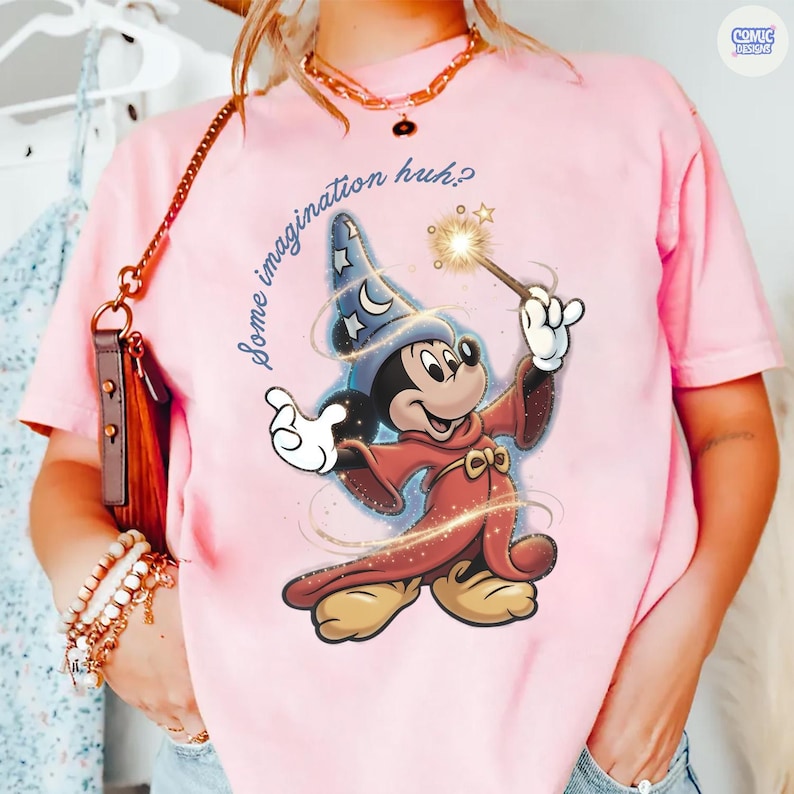 May include: Pale pink t-shirt with a graphic of Mickey Mouse as a wizard, holding a wand with a star. The text "Some imagination huh?" is above the character. The shirt has short sleeves and a round neck.