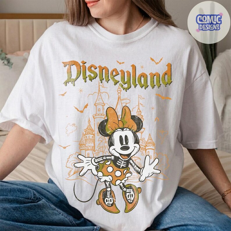 Może przedstawiać: Biały t-shirt z grafiką szkieletu Myszki Minnie w sp&oacute;dniczce w kropki i kokardzie. Nad postacią znajduje się napis Disneyland zieloną, ściekającą czcionką. Tło zawiera zarys zamku oraz latające nietoperze.