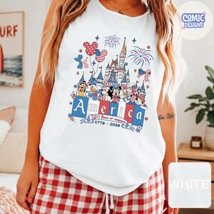 May include: A white sleeveless tank top featuring a graphic print of a castle, fireworks, and cartoon characters wearing patriotic red, white, and blue outfits. The text reads America, 250 years of freedom, and 1776-2026 in a stylised font.