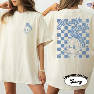 May include: Ivory t-shirt with a blue Donald Duck cartoon graphic on the front and back. The back design features the text "Donald Duck" over a blue and white chequered pattern. The shirt is labelled "Comfort Colors" and "Ivory".
