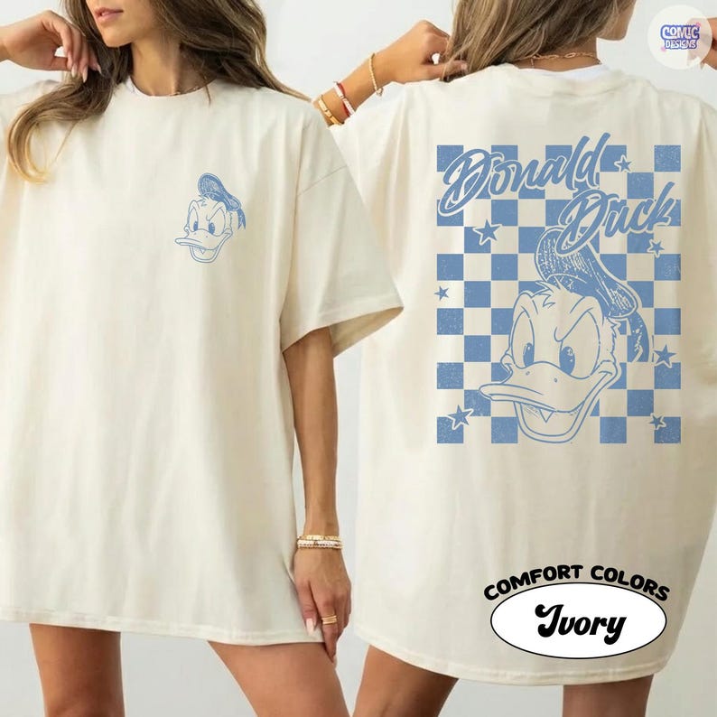May include: Ivory t-shirt with a blue Donald Duck cartoon graphic on the front and back. The back design features the text "Donald Duck" over a blue and white chequered pattern. The shirt is labelled "Comfort Colors" and "Ivory".