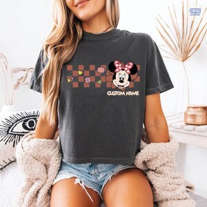 May include: Dark gray t-shirt featuring a Minnie Mouse graphic with a red bow and the text "CUSTOM NAME". The design includes a brown and pink checkered pattern with hearts and flowers. The shirt is a casual wear item.
