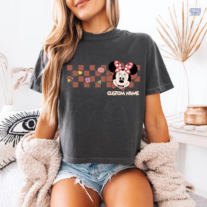 May include: Dark gray t-shirt featuring a Minnie Mouse graphic with a red bow and the text "CUSTOM NAME". The design includes a brown and pink checkered pattern with hearts and flowers. The shirt is a casual wear item.