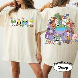 May include: An ivory t-shirt featuring a front graphic with the word NAME in colorful, patterned block letters and a back graphic displaying a purple backpack filled with various cartoon characters, including a space ranger, a cowboy, and a dinosaur.