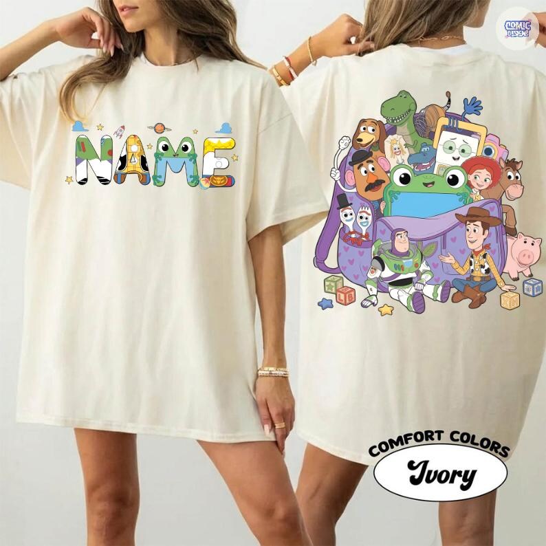 May include: An ivory t-shirt featuring a front graphic with the word NAME in colorful, patterned block letters and a back graphic displaying a purple backpack filled with various cartoon characters, including a space ranger, a cowboy, and a dinosaur.