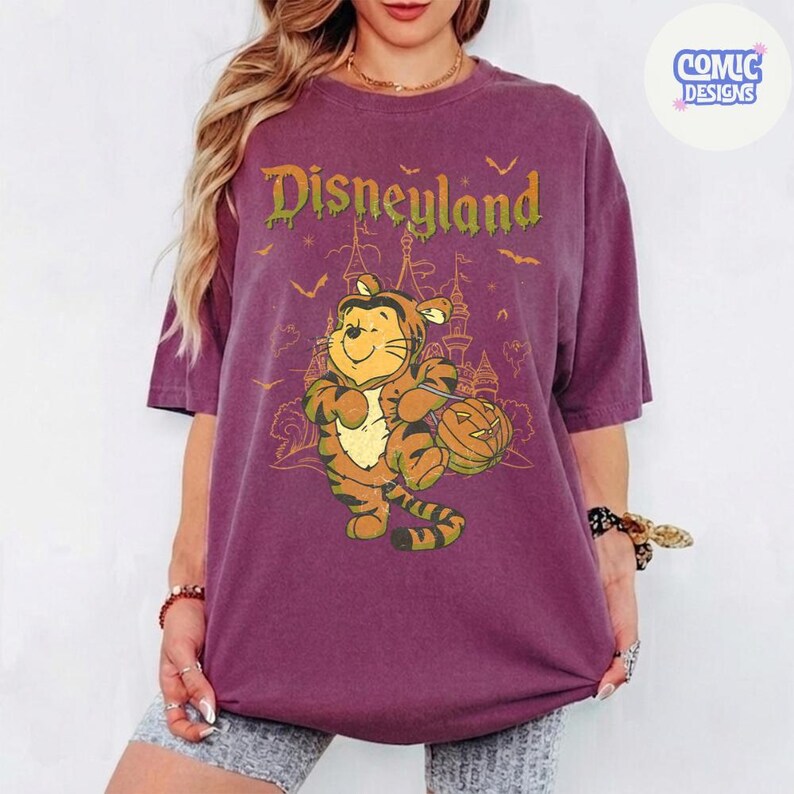 Może przedstawiać: Śliwkowy t-shirt z grafiką przedstawiającą postać przebraną za tygrysa trzymającą dynię. Napis "Disneyland" w kolorze zielonym znajduje się nad postacią, a w tle widać zarys zamku i nietoperze. Widoczne jest logo "COMIC DESIGNS".